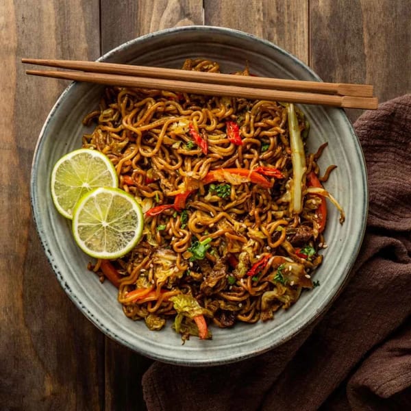 A bowl of Indian-style noodles with vegetables, lime slices, and chopsticks on a wooden table.