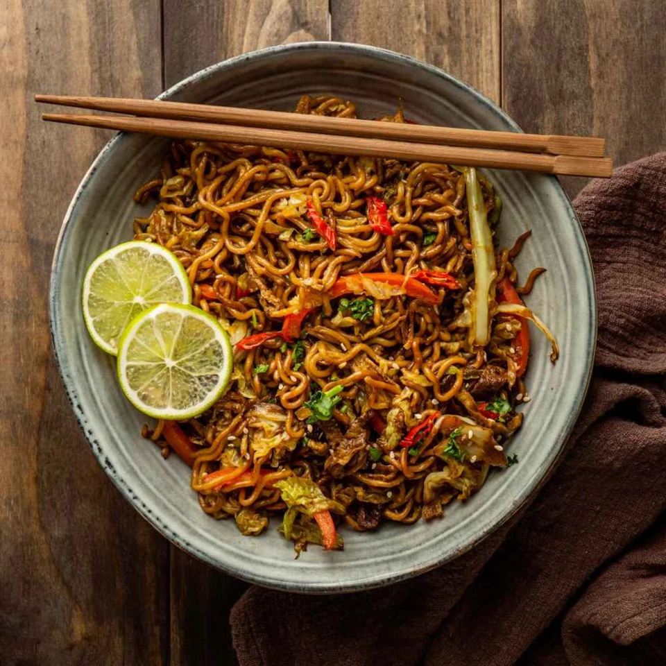 A bowl of Indian-style noodles with vegetables, lime slices, and chopsticks on a wooden table.