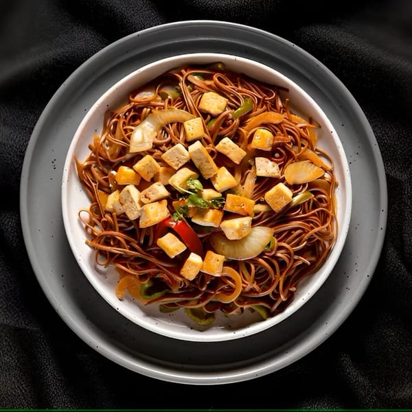 A bowl of Indian noodles with tofu, onions, and peppers on a dark background.