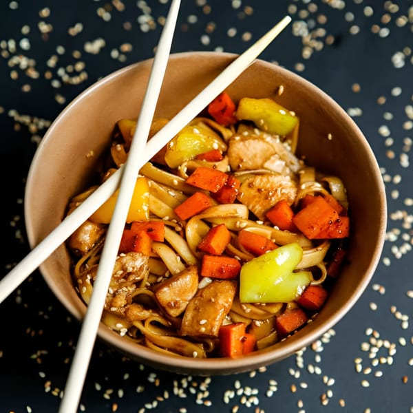 Noodles with chicken, carrots, and bell peppers in a bowl with chopsticks, sprinkled with sesame seeds.